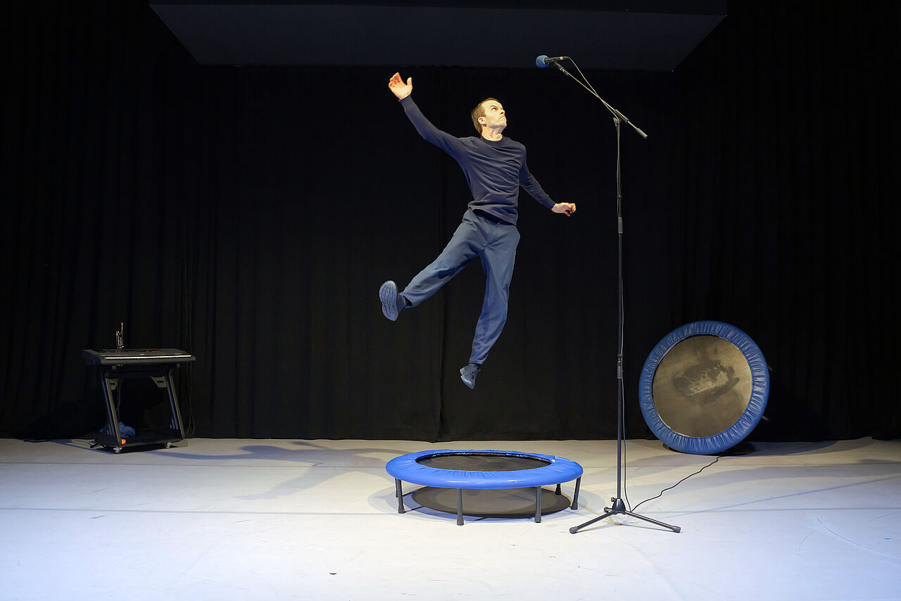 Eine Person hüpft auf einem Trampolin, um an ein viel zu hoch eingestelltes Mikrofon zu kommen und etwas zu sagen.