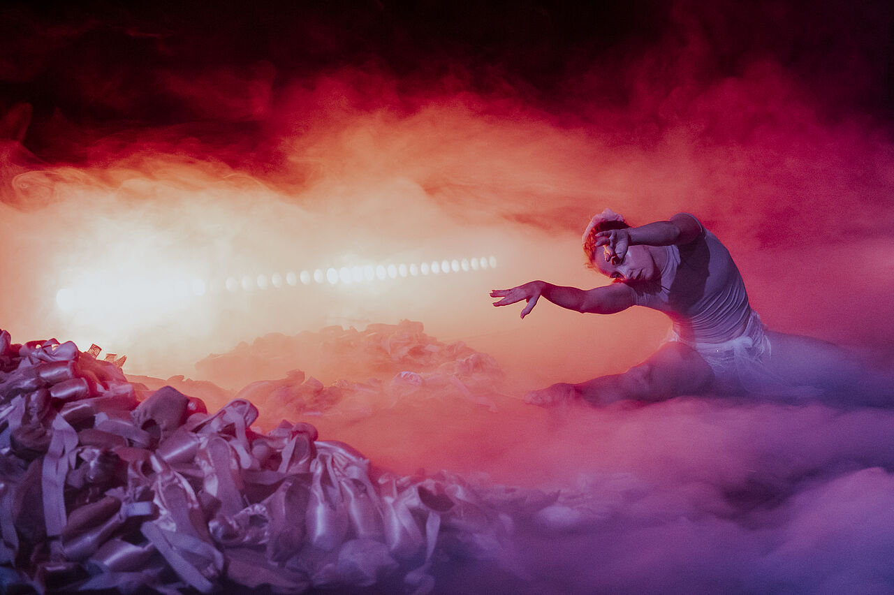 Eine Tänzerin in violettes Licht gehüllt sitzt im Spagat im Bühnenbild zwischen Nebel und hunderten von Ballettschuhen. Im Hintergrund scheint ein helles, rötlich-violettes Licht.