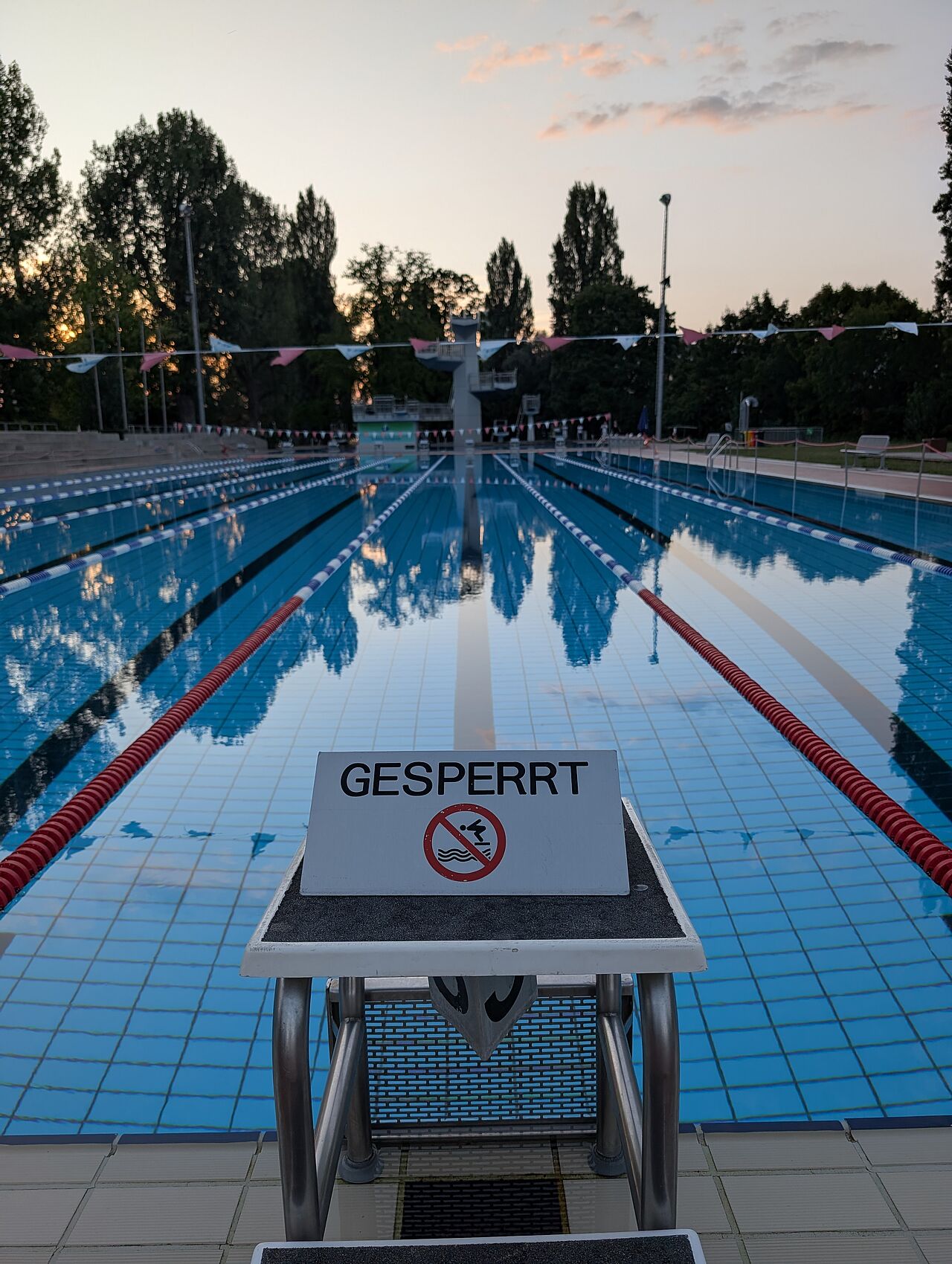 [Translate to English:] Im Hintergrund ein Schwimmenbecken im Sonnenuntergang, auf dem Startblock vor dem Becken steht ein Schild mit der Aufschrift GESPERRT.