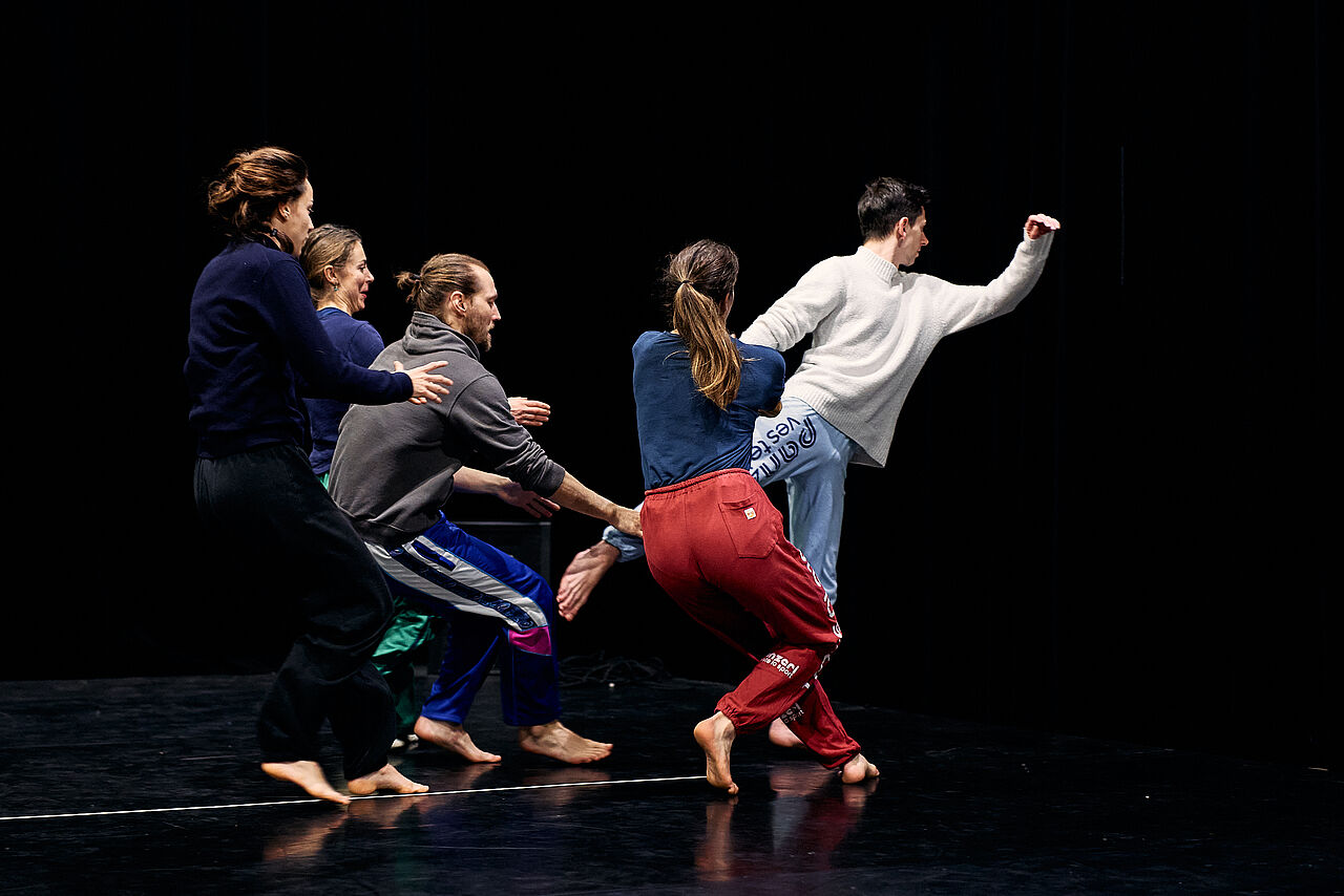 Vier Tänzer*innen halten eine fünfte Tänzer*in am Knöchel ihres linken Fuß fest. Die 5. Tänzer*in posiert in Ballett-ähnlicher Pose von den anderen weg gewandt.