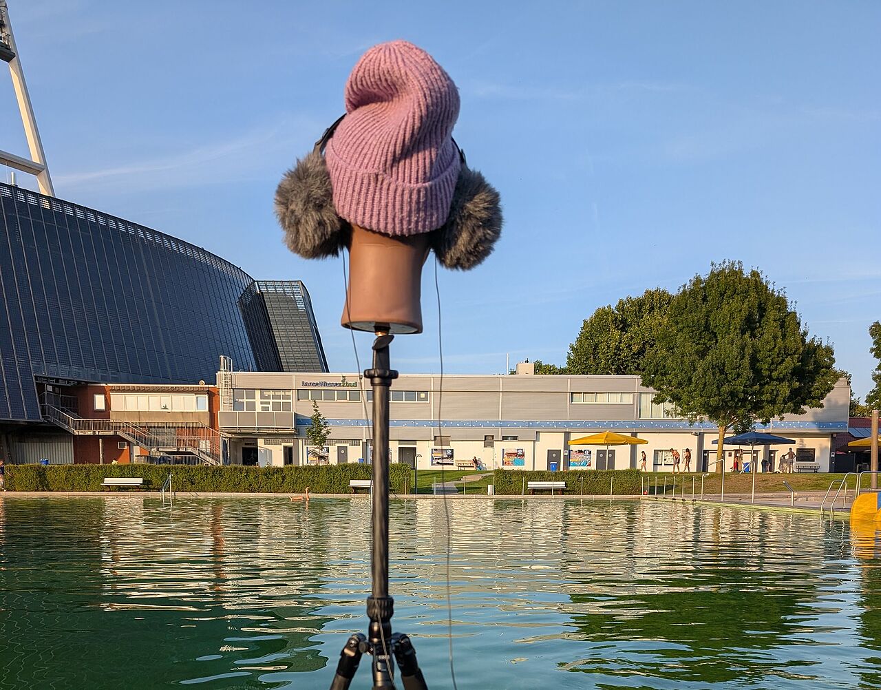[Translate to English:] Der Kopf einer Schaufenster-Puppe steht auf einem Stativ vor einem Schwimmbecken im Freibad. Er trägt eine Mütze und Kopfhörer.