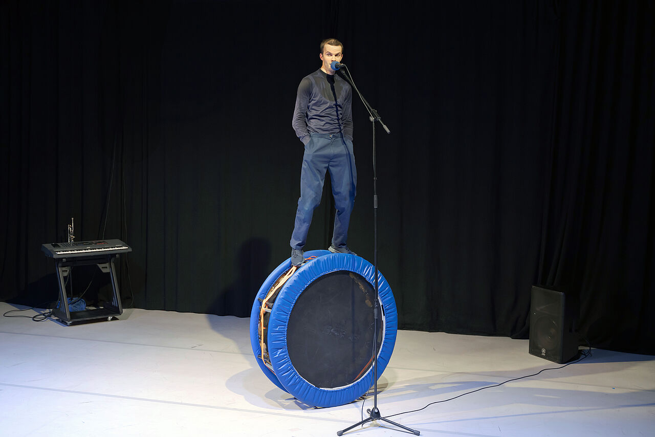 Eine Person steht auf einem runden Trampolin, was hochkant aufgestellt ist, damit sie in ein Mikrofon sprechen kann, dessen Stativ viel zu hoch eingestellt ist.