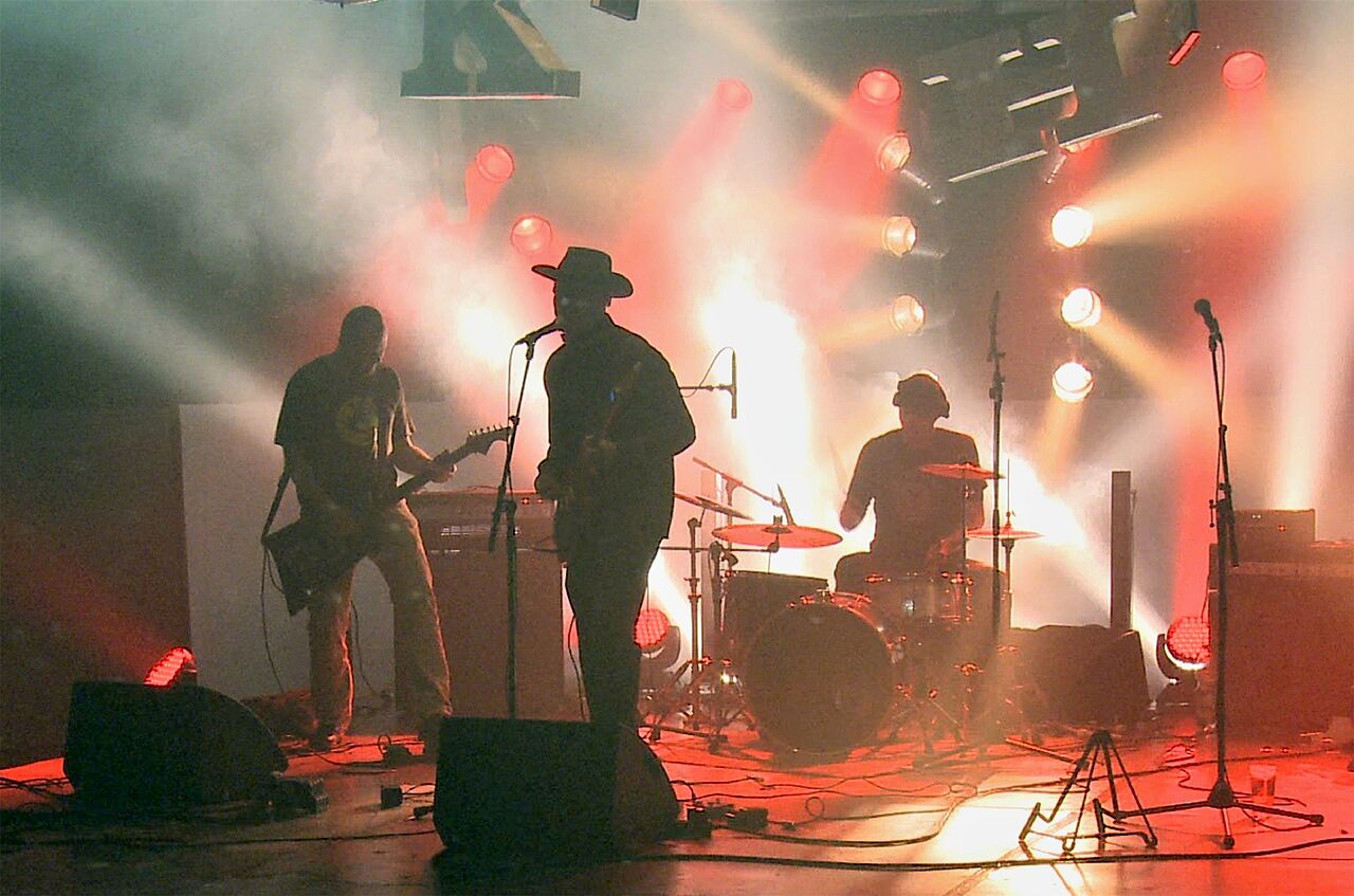 Die Schatten-Silhouetten von einer dreiköpfigen Band (Gesang, Bass/Gitarre, Schlagzeug) sind vor hellem und rotem Licht erkennbar.