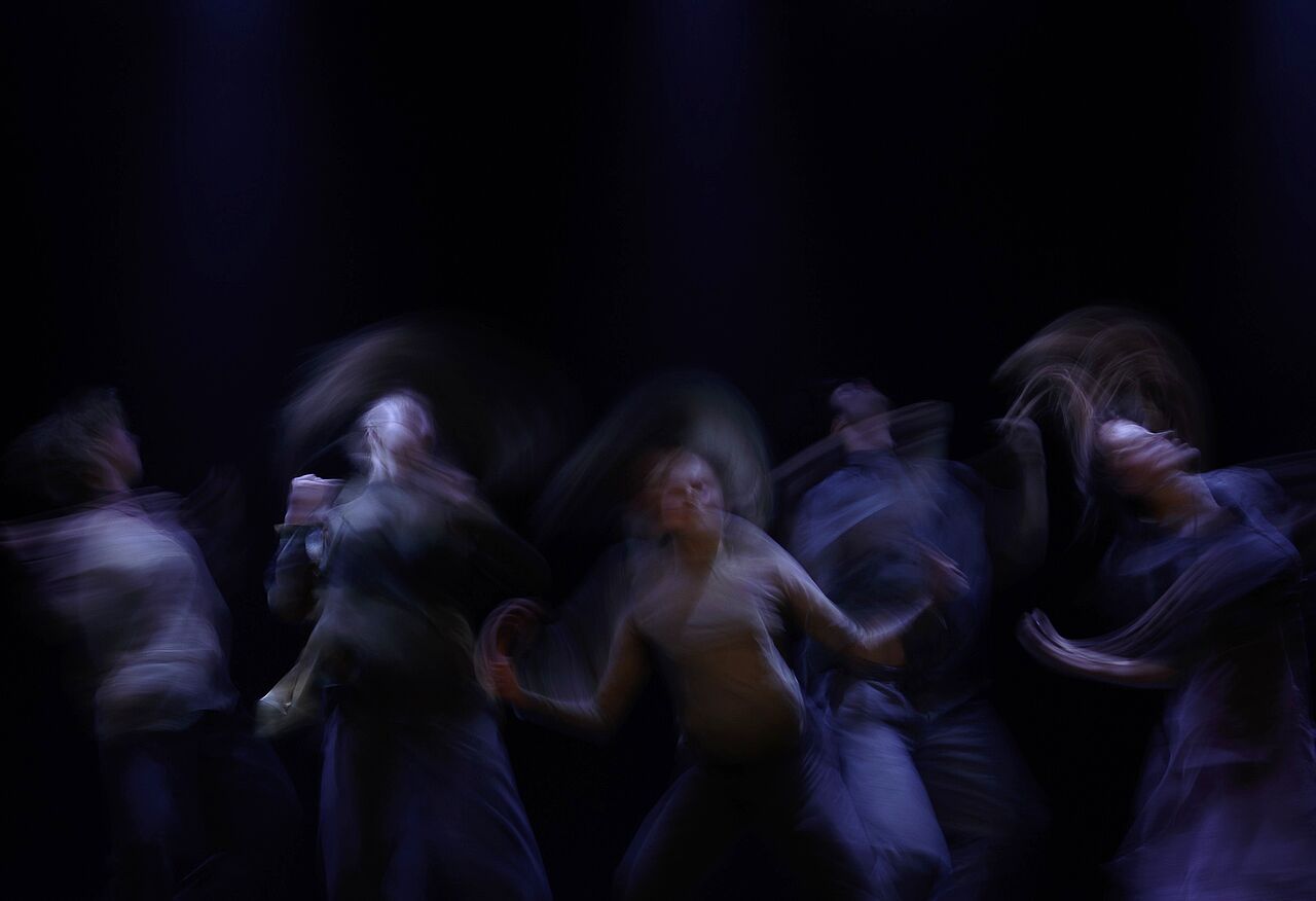 In einem dunklen Raum bewegt sich eine Gruppe von Menschen in tanzenden Bewegungen. Sie sind halb durchsichtig und ihre Bewegungenüberlagern sich, als würden sie in der Luft hängenbleiben. 