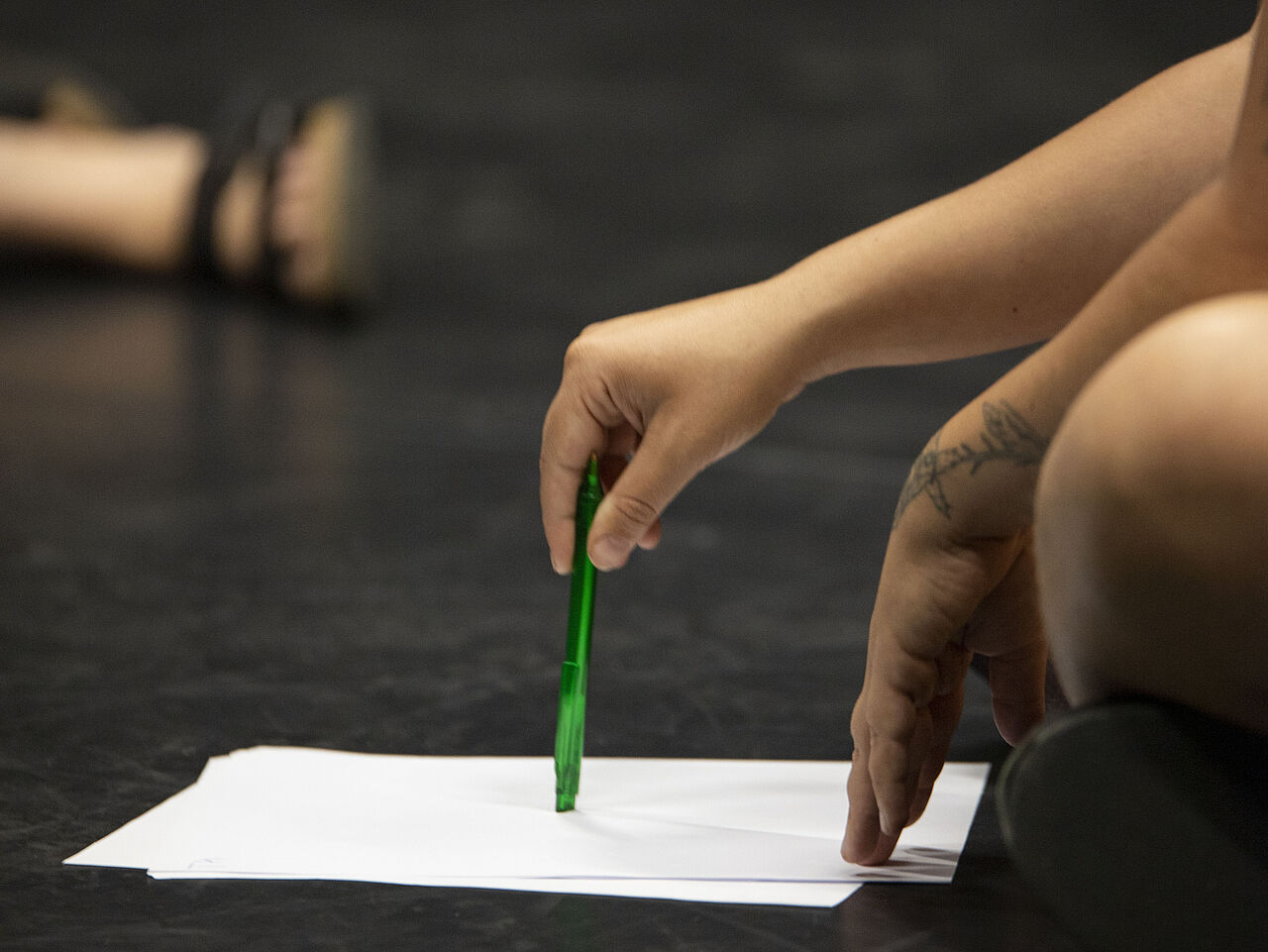 Eine Hand hält einen Stift der auf ein weißes Blatt Papier, das auf einem schwarzen Bühnenboden liegt, zeigt.
