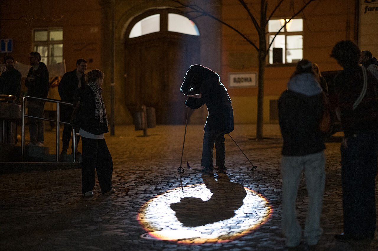 Auf einer dunklen Straße steht eine Figur auf skiern, die einen Bären-ähnlichen Kopf hat und Anzug trägt. Sie wird von einem Lichtspot beleuchtet.